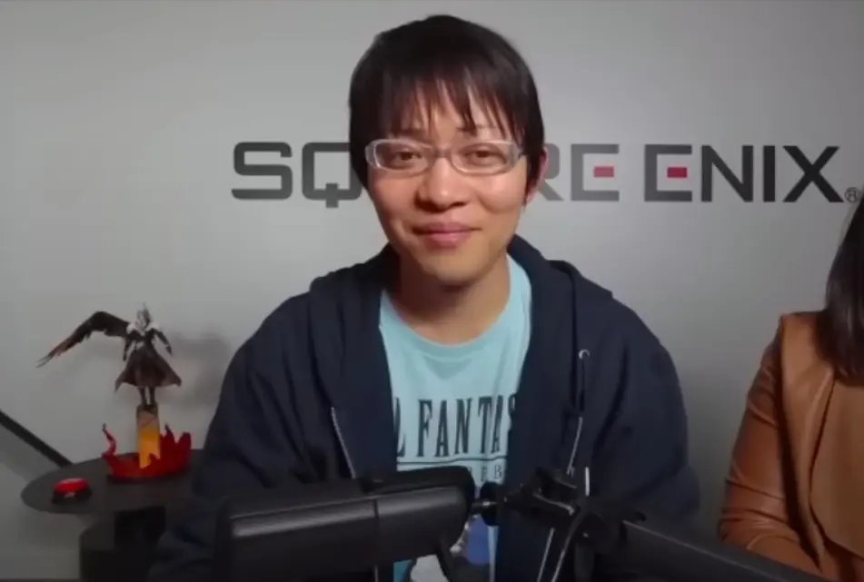 Si Naoki Hamaguchi na nakasuot ng salamin at asul na t-shirt, habang nakaupo sa harap ng isang Square Enix wall.