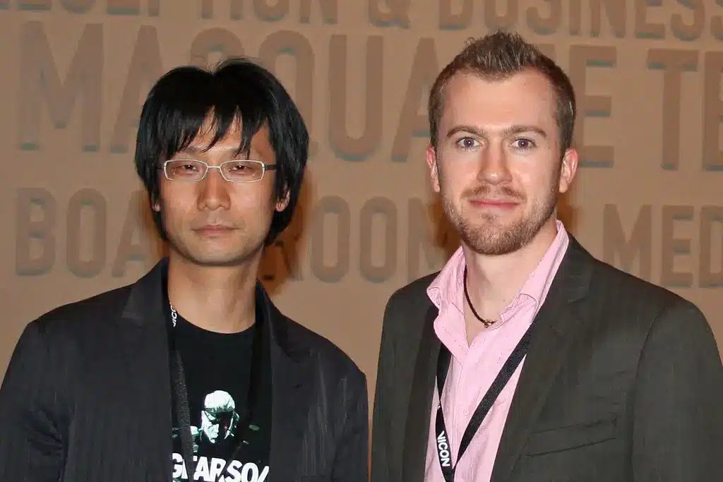 Si Hideo Kojima na nakasuot ng salamin at itim na blazer, habang nakatayo sa tabi ng isa pang lalaki.
