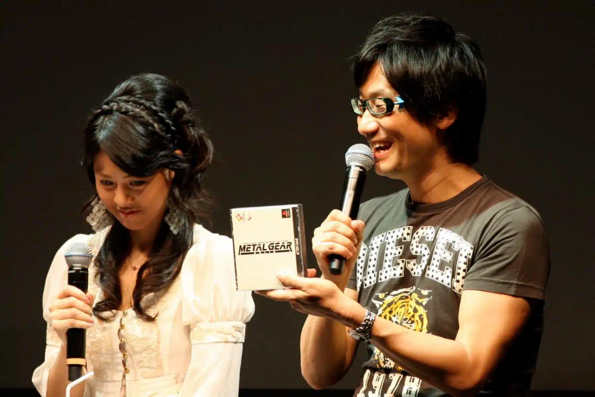 Si Hideo Kojima na may hawak na case ng Metal Gear habang nagsasalita sa harap ng mikropono.